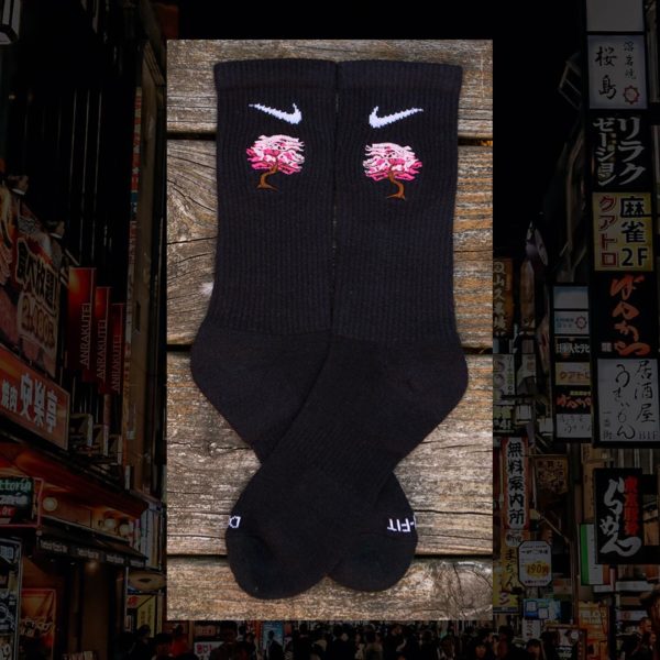 Steinblokk - Black crew sock with a pink and brown embroidery of a cherry blossom tree. The photo was taken under the glow of sunset with a wood background.