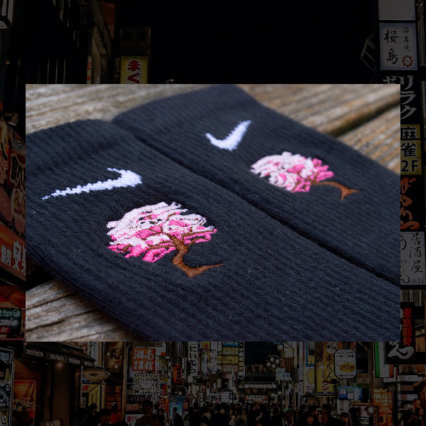 Steinblokk - Black crew sock with a pink and brown embroidery of a cherry blossom tree. The photo was taken under the glow of sunset with a wood background.