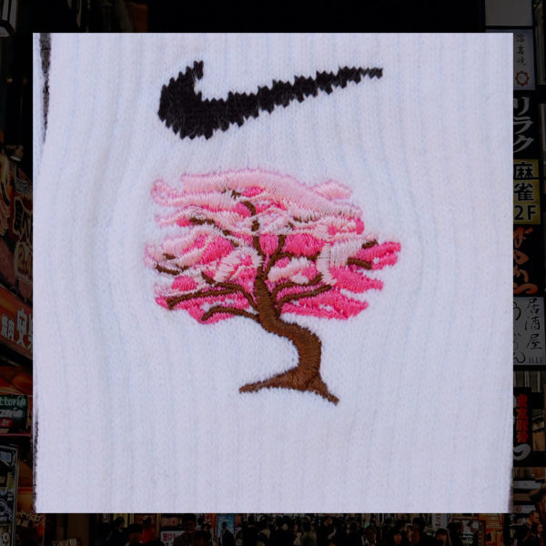 Steinblokk - White crew sock with a pink and brown embroidery of a cherry blossom tree. The photo was taken under the glow of sunset with a wood background.