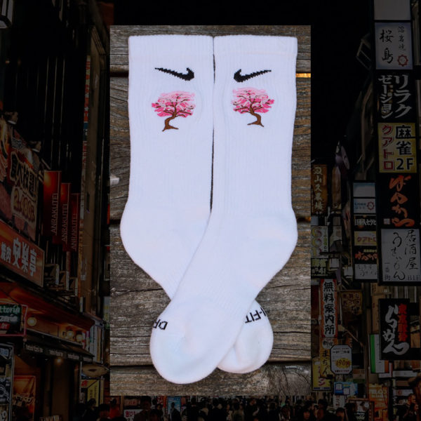 Steinblokk - White crew sock with a pink and brown embroidery of a cherry blossom tree. The photo was taken under the glow of sunset with a wood background.