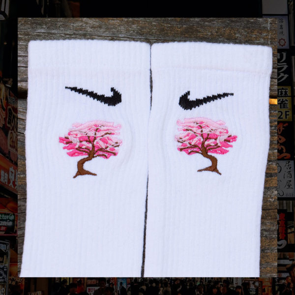 Steinblokk - White crew sock with a pink and brown embroidery of a cherry blossom tree. The photo was taken under the glow of sunset with a wood background.