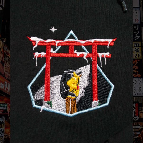 Steinblokk - Embroidery depicting a mountaineer hiking up Mt. Fuji at night. The climber is passing under a tori gate covered in ice. The climber wears a yellow jacket, brown pants, and a black backpack. The design is bordered by ice blue stitching.